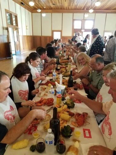 Happy guests enjoying lobster feed, tables filled with bread, wine, lobster, lemons and artichokes