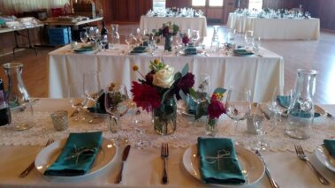 White table tops with shiny glassware, green fabric napkins and fresh cut white roses and red dahlia flower arrangement