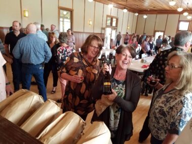 3 women laughing and holding bottle of wine at Lobster Feed, room full of people in background