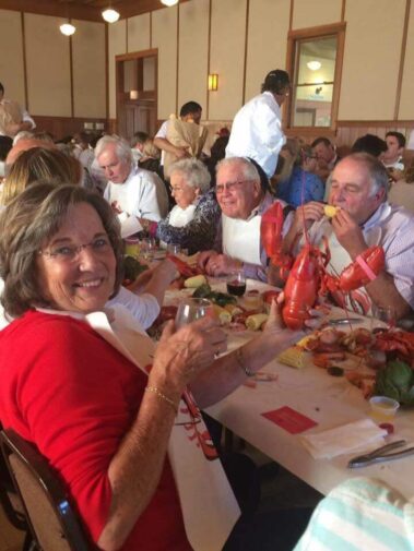 Smiling guests enjoying lobster dinner at table with lobster in one woman's hand and glass of wine in the other