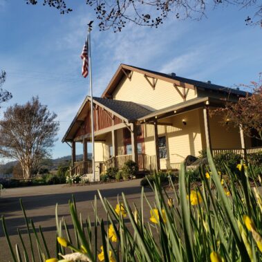 Front view of yellow and brown building with front porch and American flag. Yellow daffodils in foreground.